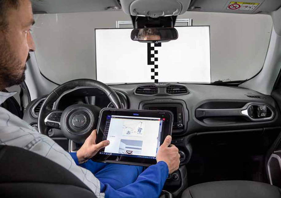 Automobilio salone sėdintis technikas su diagnostine planšete atlieka ADAS kameros kalibravimą priešais specialų kalibravimo ekraną.