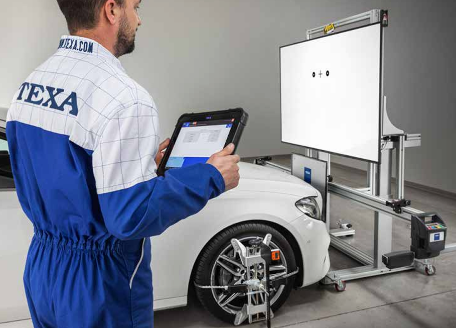 ADAS kalibravimo stendas automobilių servise, technikas su planšete kalibruoja automobilio pagalbos vairuotojui sistemas naudojant ratų suvedimo įrangą.