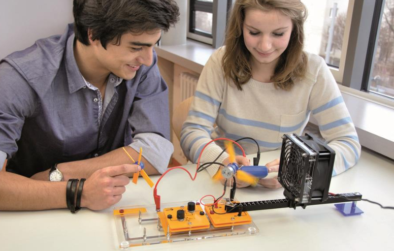 Studentai atlieka vėjo energijos laboratorinį eksperimentą su ventiliatoriumi ir turbinos modeliu, matuojant pagamintą elektros energiją.