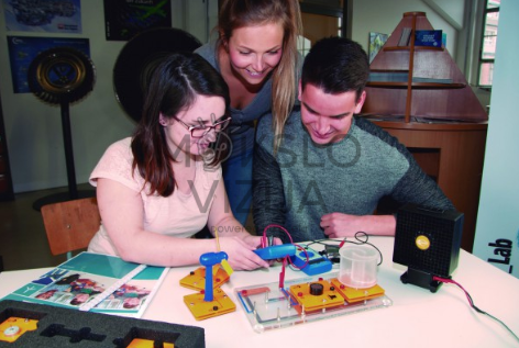 Trys studentai atlieka vėjo energijos eksperimentą su mokomąja turbina ir laboratorine įranga.