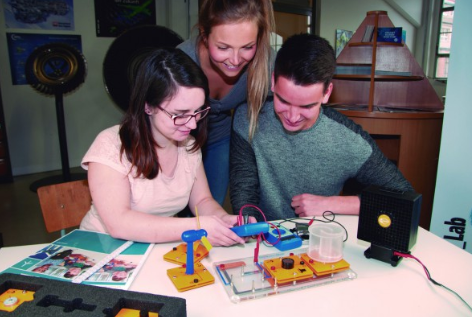 Trys studentai atlieka vėjo energijos eksperimentą su mokomąja turbina ir laboratorine įranga.