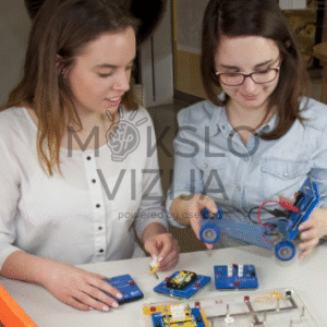 Dvi studentės laboratorijoje atlieka elektromobilumo eksperimentą su elektrinio automobilio modeliu ir moduliniais komponentais.