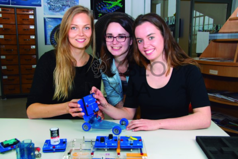 Trys studentės laboratorijoje demonstruoja elektromobilumo eksperimentą su elektrinio automobilio modeliu ir energijos kaupimo moduliais.