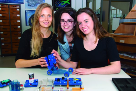 Trys studentės laboratorijoje demonstruoja elektromobilumo eksperimentą su elektrinio automobilio modeliu ir energijos kaupimo moduliais.