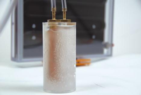 Vandenilio kuro elemento cilindrinis modulis su burbuliuojančiu skysčiu ir prijungtais vamzdeliais laboratoriniam eksperimentui.