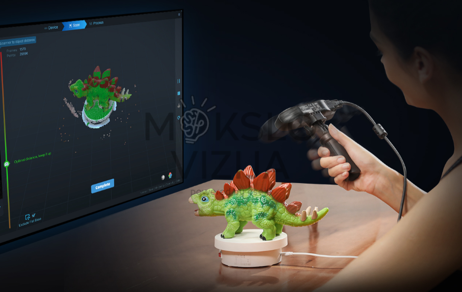 Asmuo rankiniu 3D skeneriu skenuoja žaislinį modelį ant stalo, o monitoriuje rodomas spalvotas 3D objekto modelis.