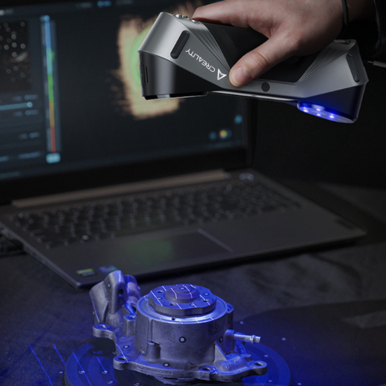 Rankinis 3D skeneris su mėlyno lazerio apšvietimu skenuoja metalinę detalę ant stalo, šalia matomas kompiuteris su 3D skenavimo programa.