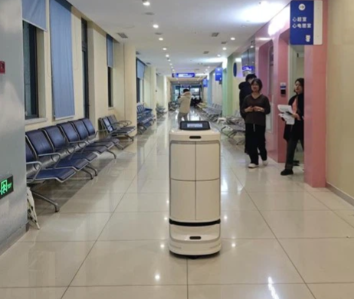 Autonominis pristatymo robotas Speedybot važiuoja ligoninės koridoriumi tarp laukiamojo zonos kėdžių.