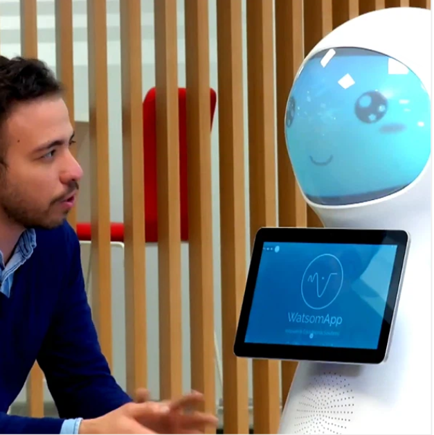 AI serviso robotas su lietimui jautriu ekranu ir animuotu veidu kalbasi su žmogumi modernioje aplinkoje.