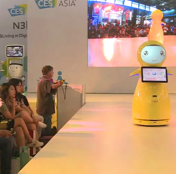 Geltonas interaktyvus AI robotas su ekranu scenoje parodos metu, skirtas lankytojų informavimui ir komunikacijai.