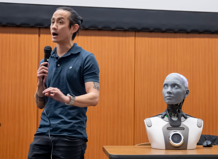 Pranešėjas su mikrofonu stovi šalia stalinio humanoidinio roboto Ameca Desktop, kuris pastatytas ant stalo konferencijų salėje ir žiūri į žmogų.