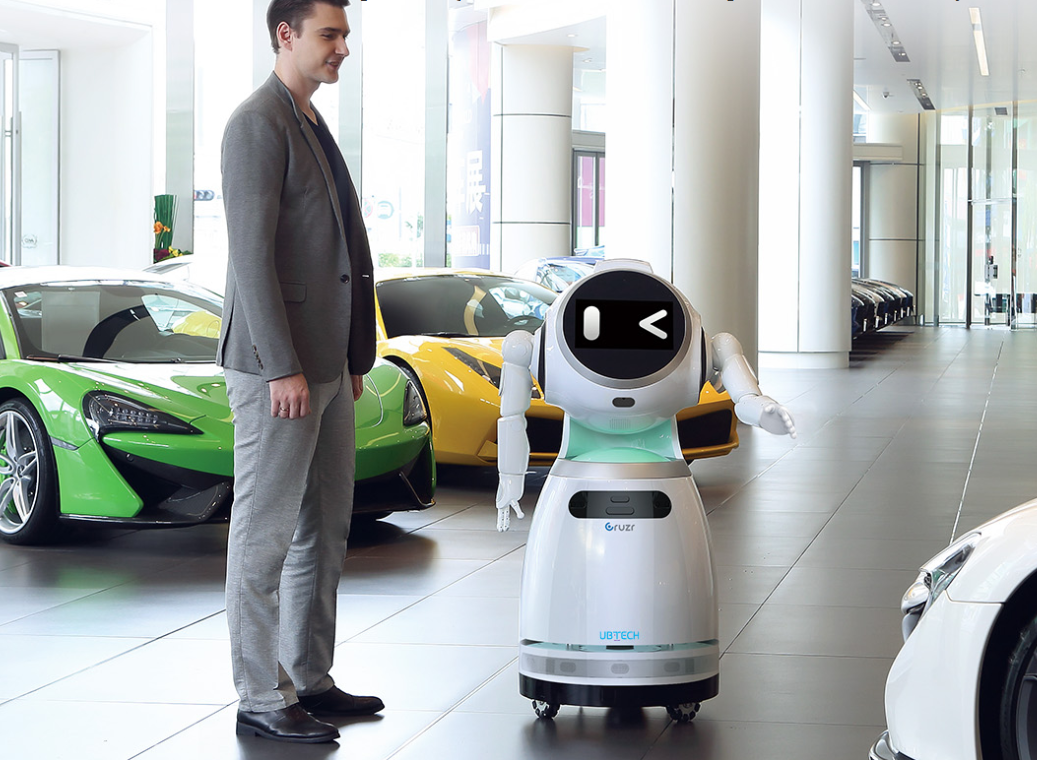 Baltas humanoidinis robotas su ekrano veidu stovi automobilių salone šalia vyro ir prabangių sportinių automobilių, demonstruodamas klientų aptarnavimo funkcijas.