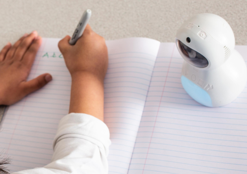 Vaiko ranka rašo sąsiuvinyje, o šalia pastatytas mažas baltas edukacinis robotas su skaitmeniniu veidu ir šviečiančia mėlyna dalimi.