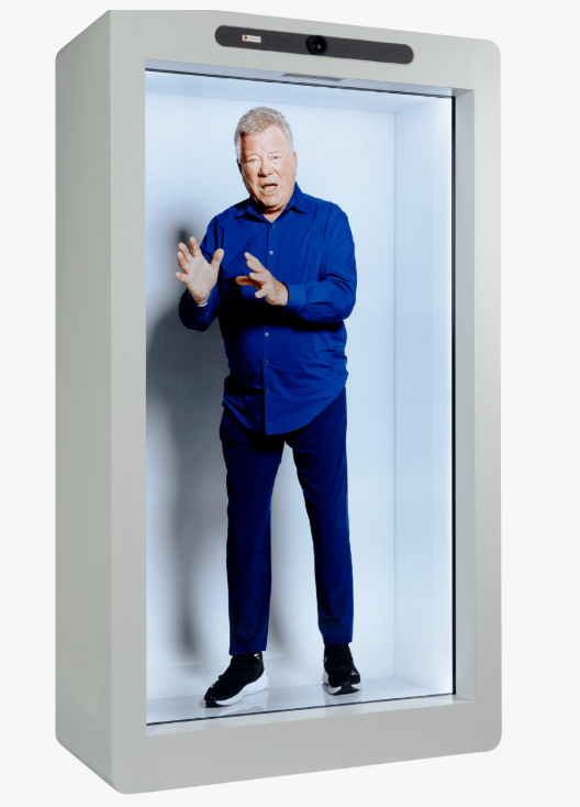 Stačiakampė holografinė projekcijos sistema su dideliu ekranu, kuriame matomas viso ūgio vyras mėlynais drabužiais baltame fone.