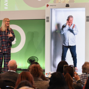 Konferencijos scenoje stovi holografinė kabina, kurioje rodomas viso ūgio vyras, o šalia scenos kalba moteris prieš auditoriją.