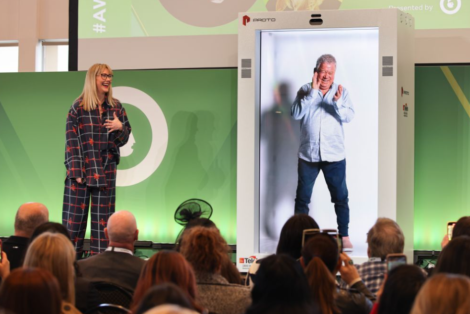 Konferencijos scenoje stovi holografinė kabina, kurioje rodomas viso ūgio vyras, o šalia scenos kalba moteris prieš auditoriją.