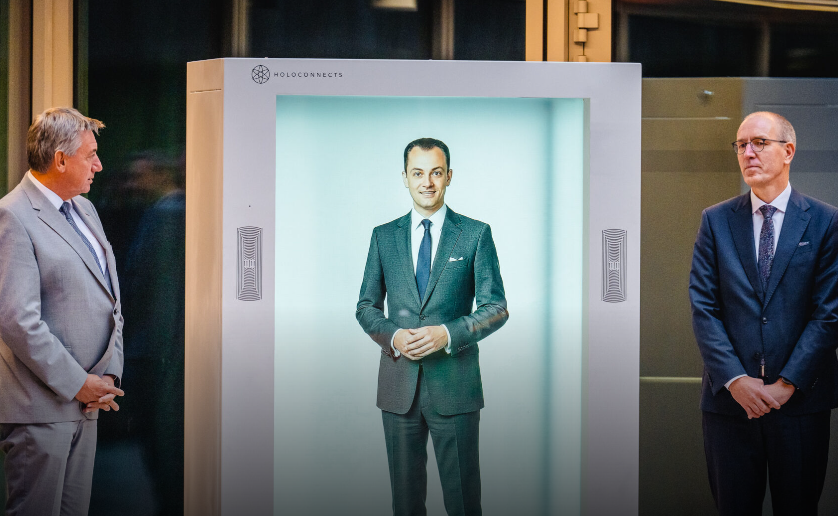 Stačiakampėje holografinėje kabinoje matomas kostiumu vilkintis vyras, o šalia stovi du vyrai verslo aplinkoje.