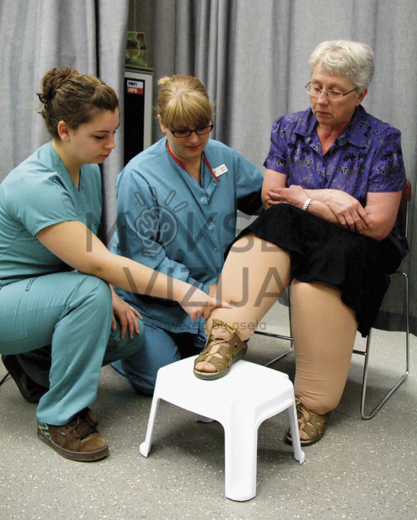 Kineziterapijos specialistės apžiūri vyresnės moters koją klinikinėje aplinkoje, vertindamos judėjimą ir galimą skausmą